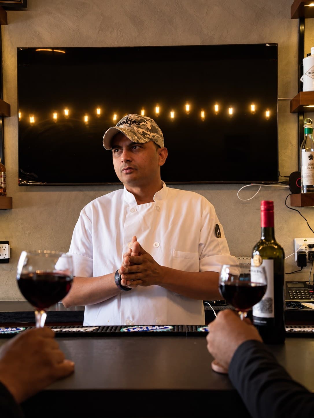 Chef Amrit Pal Singh behind the bar at Angel, hands together, glasses of red wine and a bottle in the foreground