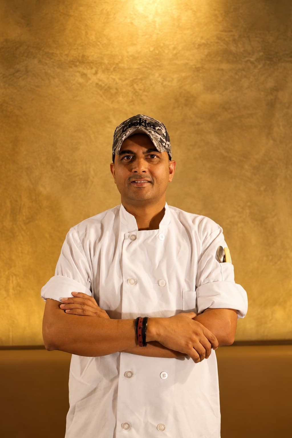 Chef Amrit Pal Singh in chef's whites and a cap, arms folded, against a warm gold wall