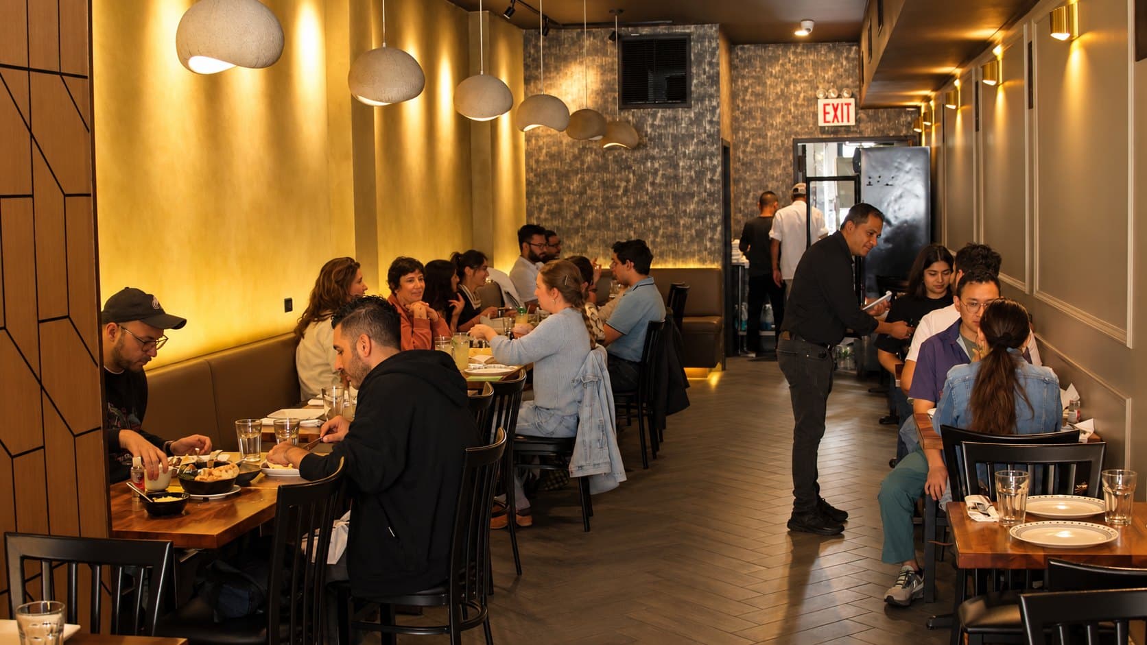 A full dining room at Angel: guests at tables along a banquette under pendant lights, a server taking an order
