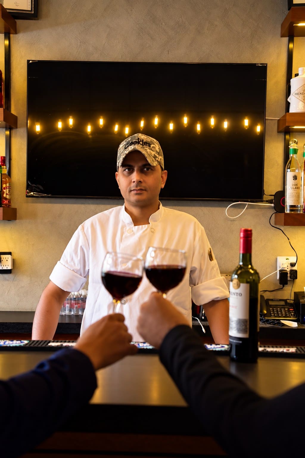 Chef Amrit Pal Singh behind the bar at Angel in chef's whites and a cap, two guests raising glasses of red wine in the foreground