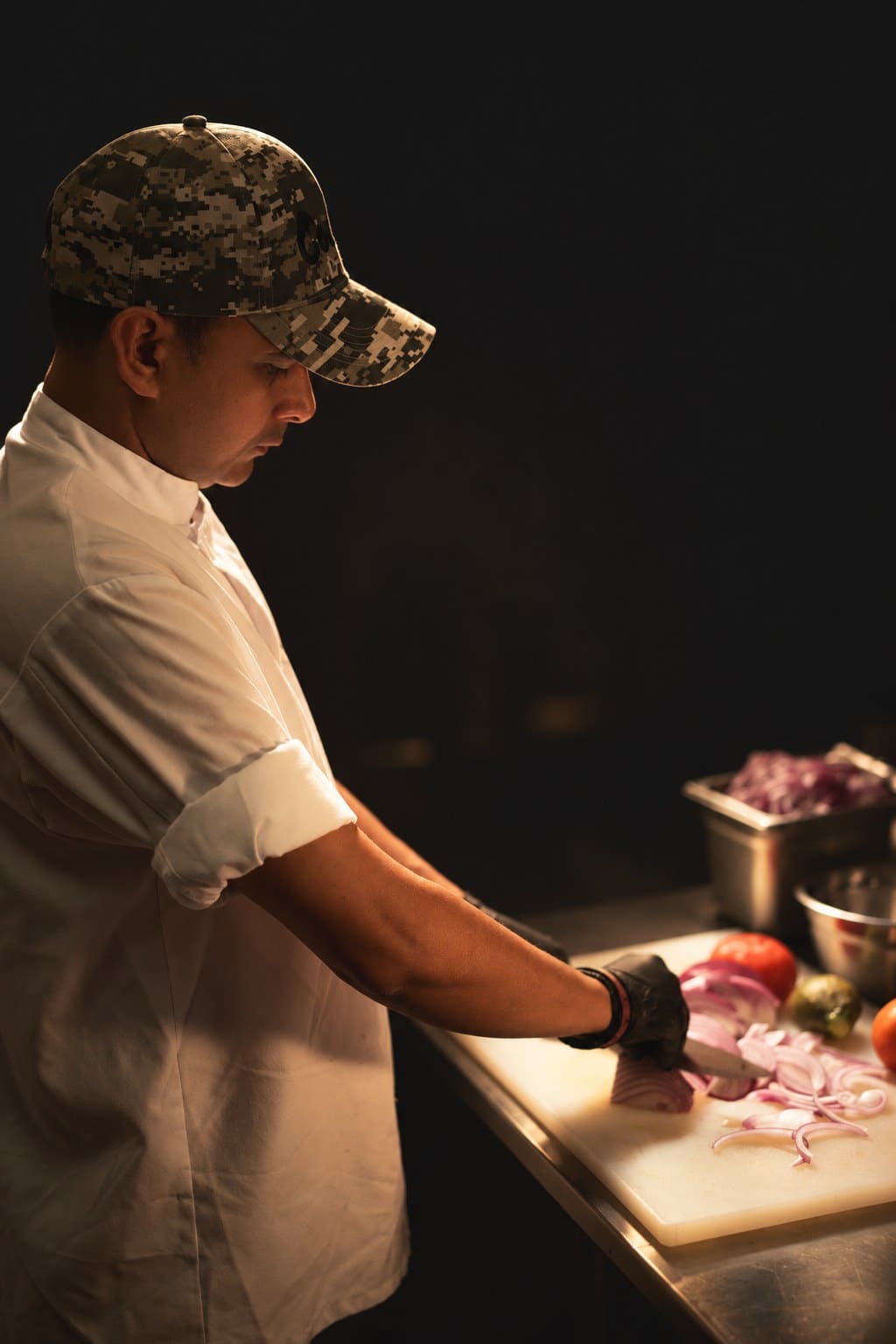 Chef Amrit Pal Singh in a cap and whites, slicing red onions on a board in a darkened kitchen