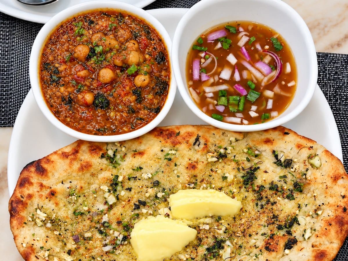 Amritsari aloo kulcha on a white plate, flecked with herbs and topped with two pats of butter, beside a bowl of chickpea curry and a bowl of onion and tomato in tangy water