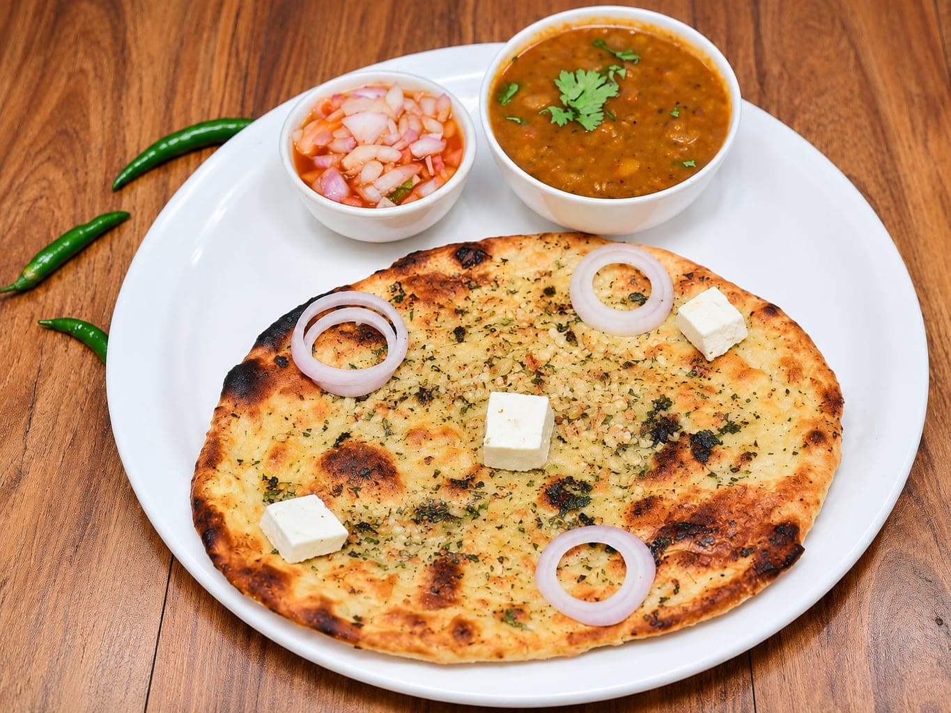 Amritsari paneer kulcha on a white plate, topped with cubes of paneer and rings of onion, with a bowl of chickpea curry, a bowl of pickled onion and green chillies on a wooden table