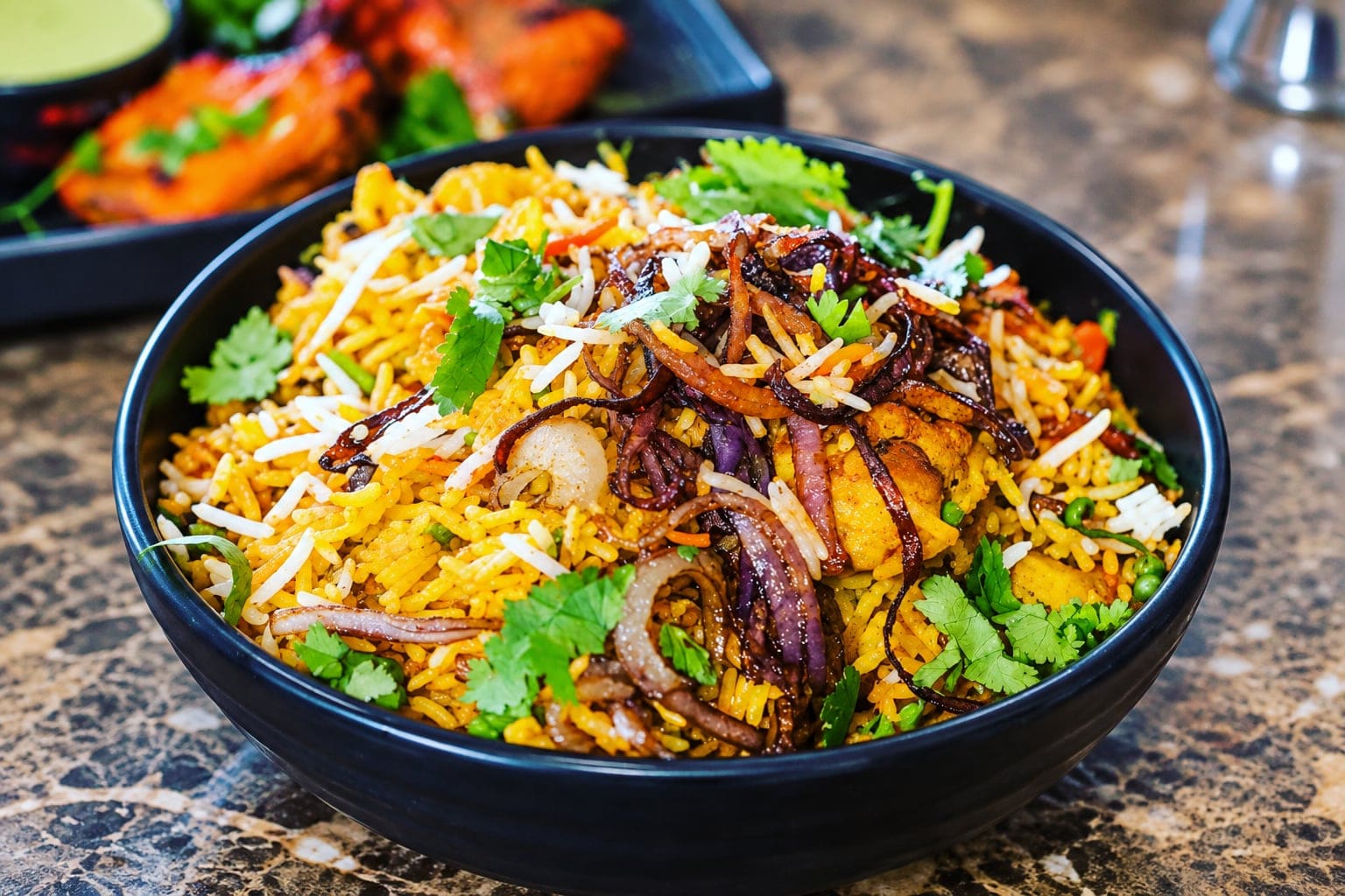 A dark bowl of chicken dum biryani: saffron basmati with pieces of chicken, crisp fried onions and fresh coriander, on a marble table