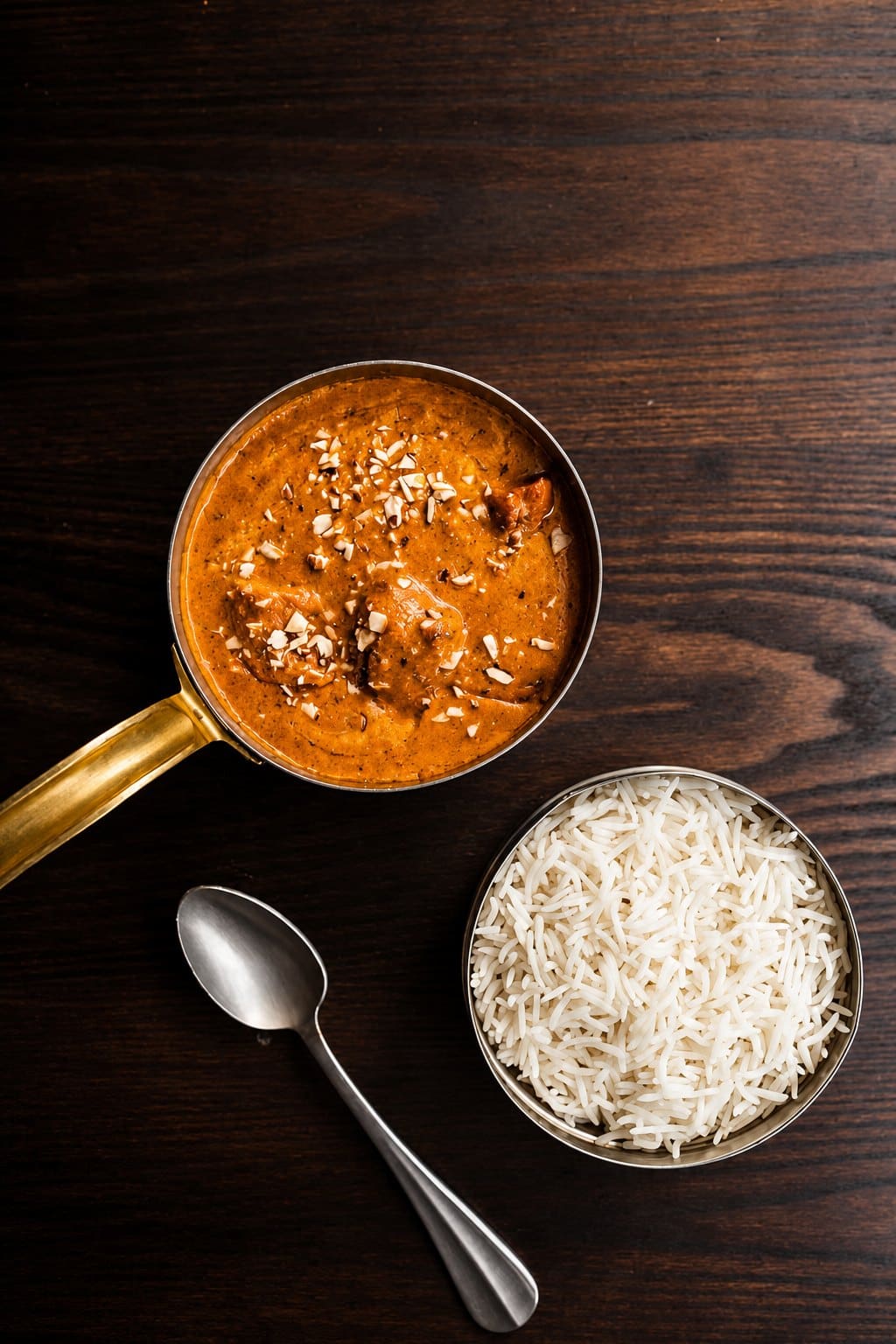 A curry topped with almonds in a copper pan, with a bowl of basmati rice on a dark wooden table