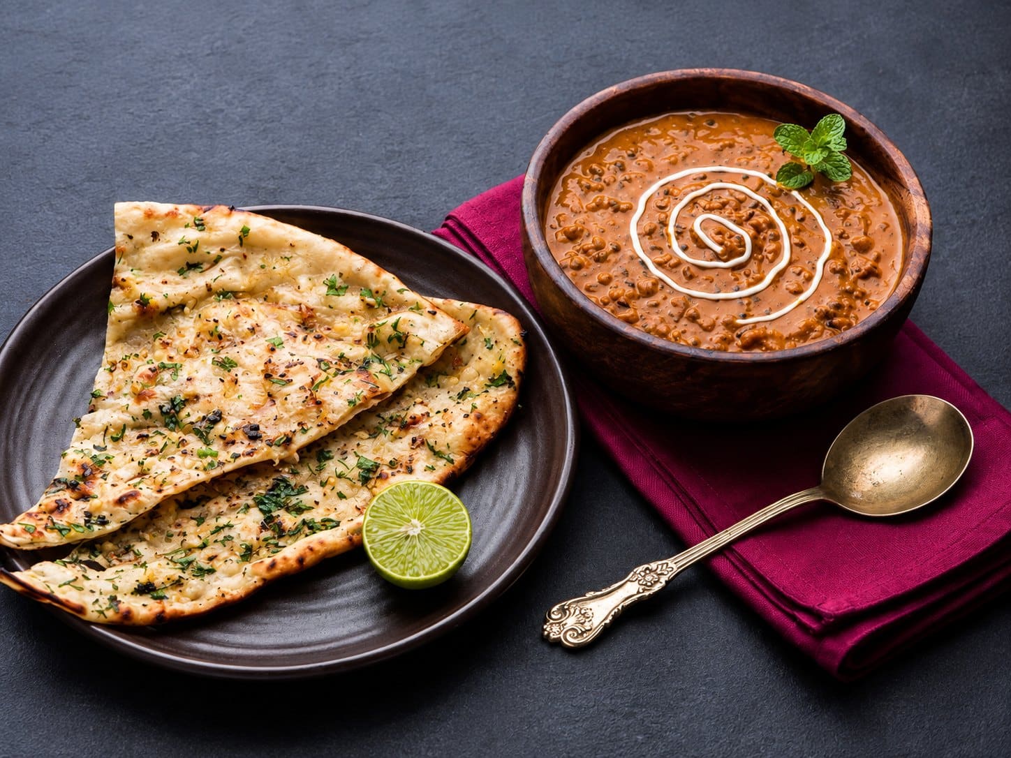 A bowl of dal makhani swirled with cream, beside garlic naan and a wedge of lime