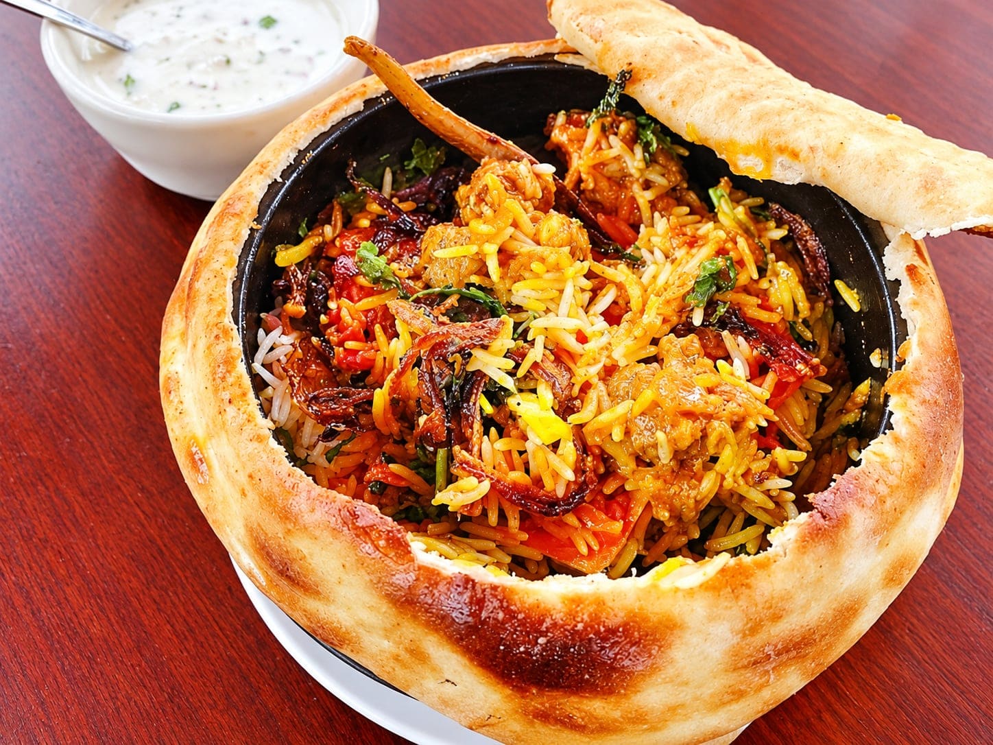 Goat dum biryani in a pot sealed with baked bread, the crust lifted to show saffron rice, goat and fried onions, with a bowl of raita, on a red wood table