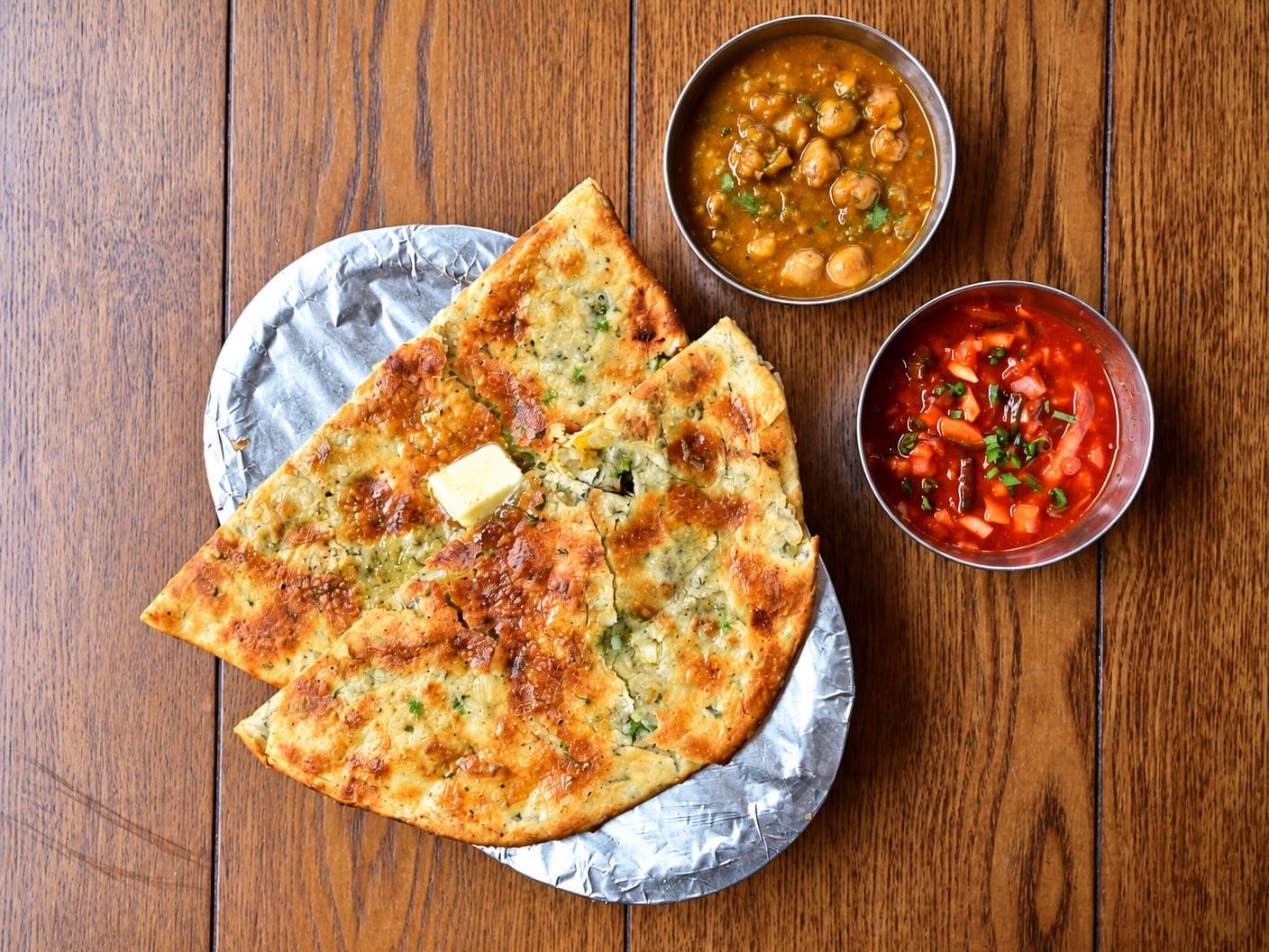 Mix veg kulcha cut into wedges on a foil-lined plate, flecked with herbs and topped with a pat of butter, beside a bowl of chickpea curry and a bowl of chopped onion and tomato, on a wooden table