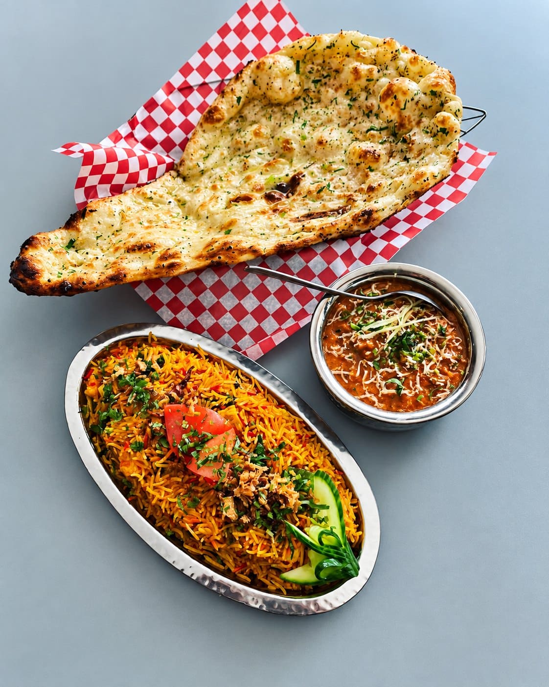 Garlic naan on red-checked paper in a basket, a platter of biryani with tomato and cucumber, and a bowl of curry