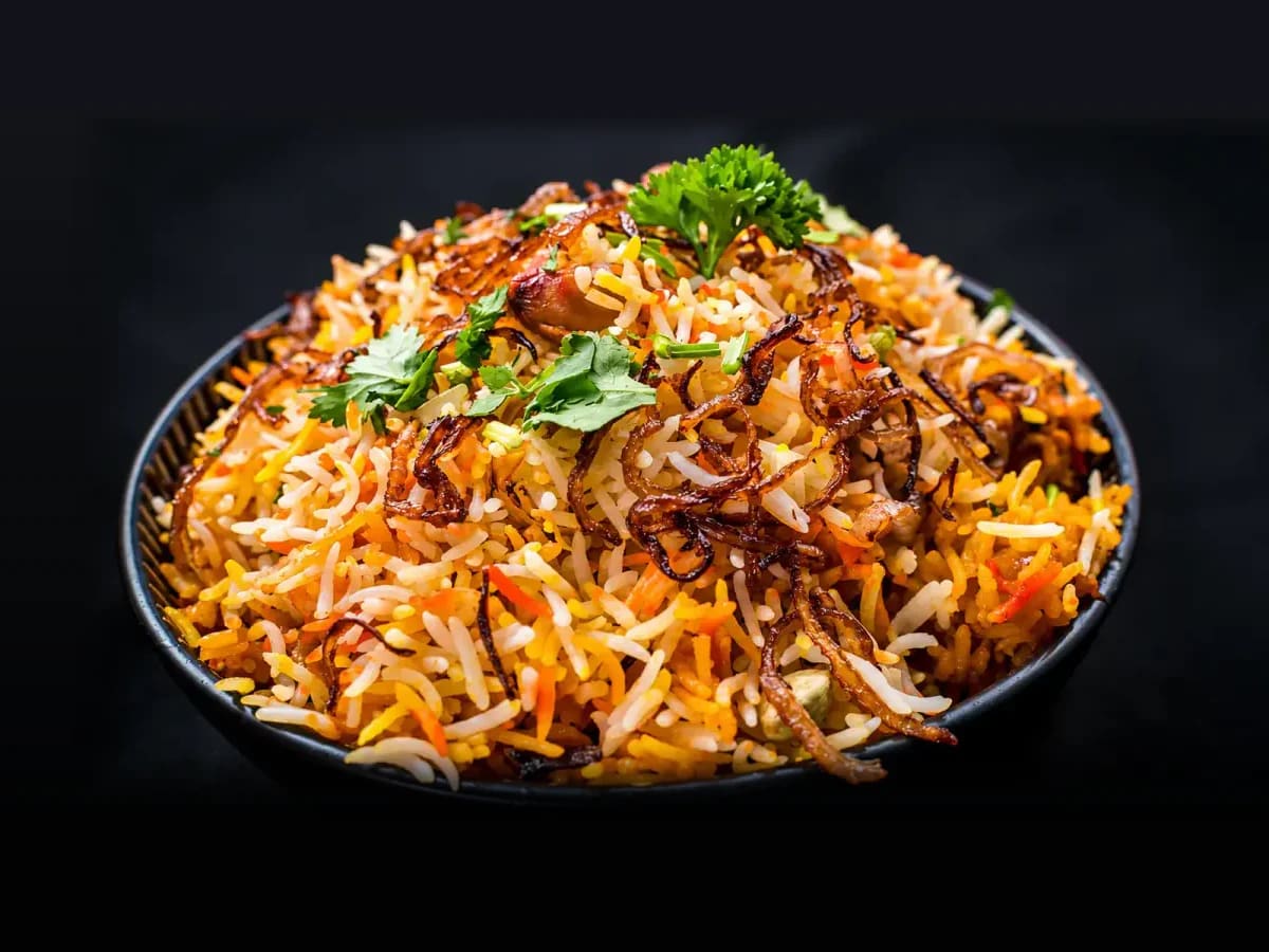 A dark bowl of vegetable dum biryani: saffron-gold basmati with carrots, crisp fried onions and fresh coriander on top