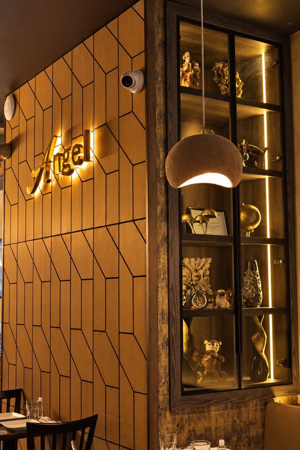 Inside Angel: the gold Angel sign on a wall of honey-oak geometric panels, a stone pendant lamp and a lit display cabinet of brass ornaments