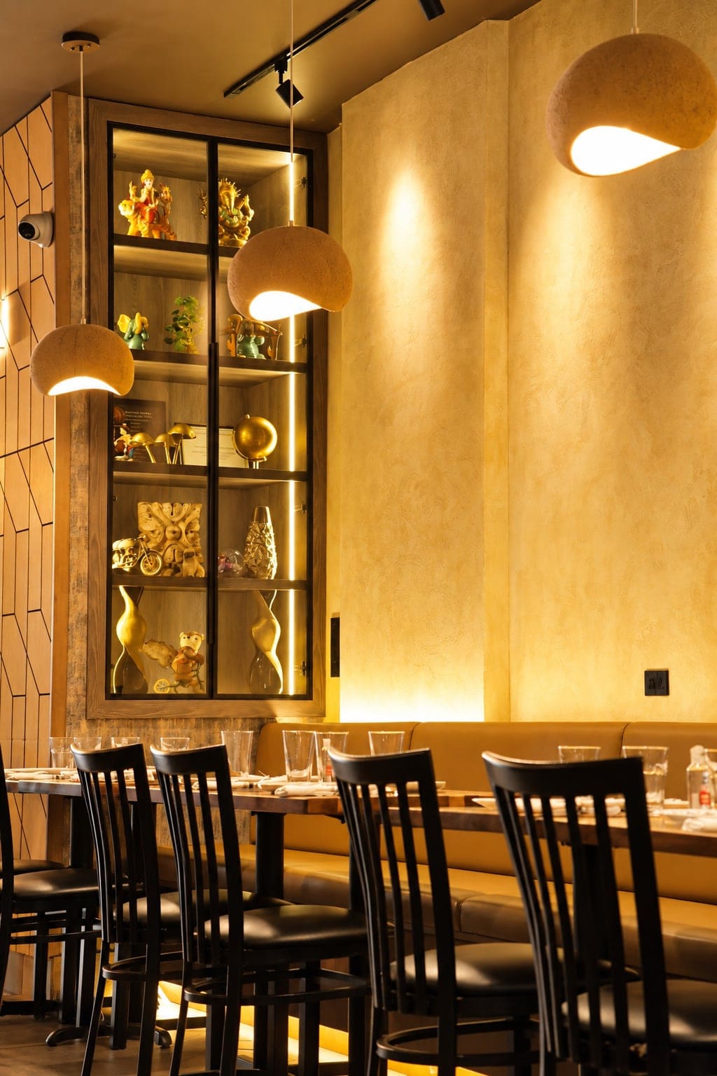 Angel's dining room in warm light: a glass display case of gold figurines, round pendant lamps, and black-backed chairs along a tan leather banquette